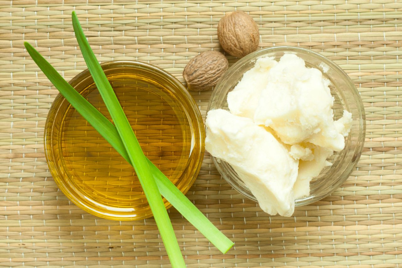 Óleo vegetal em recipiente de vidro ao lado de manteiga vegetal e folhas verdes sobre esteira natural