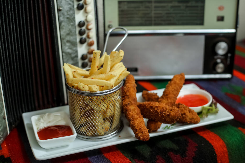 Petiscos fritos crocantes preparados em fritadeira elétrica industrial servidos com ketchup e molho picante.