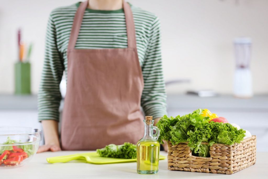 Uma pessoa de avental rosa e blusa listrada está em uma cozinha clara. À frente dela, sobre uma mesa branca, há uma pequena garrafa de vidro com óleo dourado, uma cesta de palha com alface fresca, tomates e pimentões, e uma tigela de vidro com salada picada. O fundo está levemente desfocado, sugerindo um ambiente doméstico e saudável.