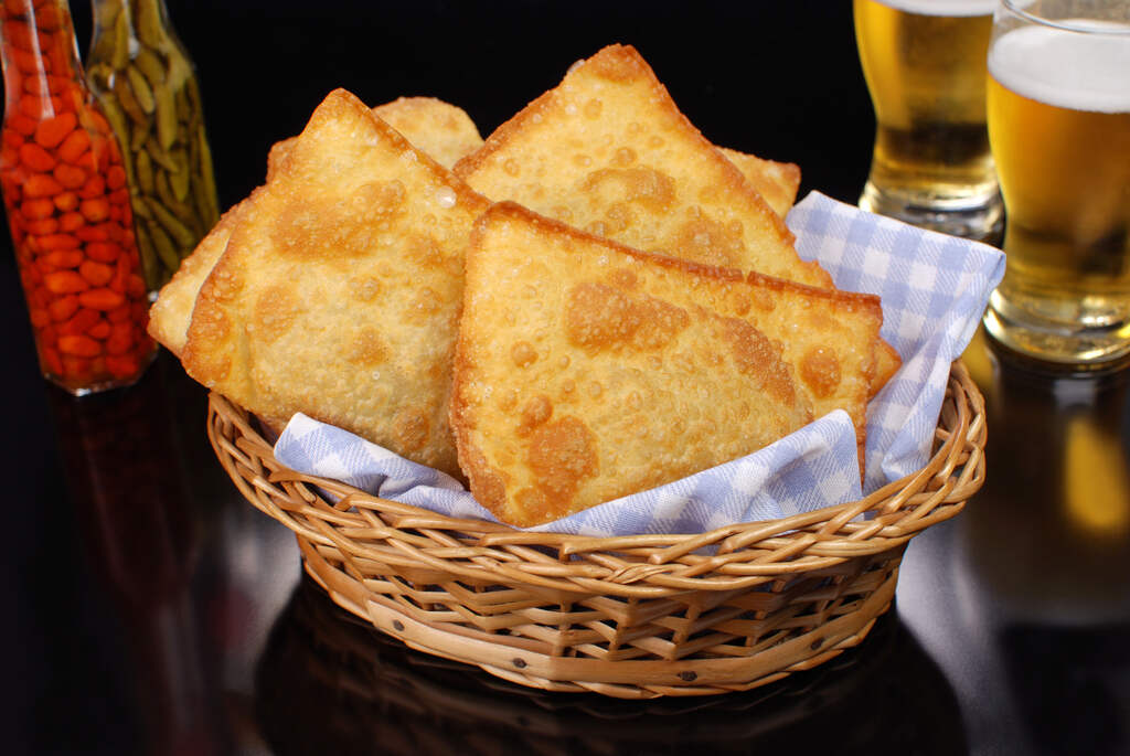 Cesta de vime com mini pastéis fritos bem dourados sobre um guardanapo xadrez azul. Ao fundo, copos de cerveja e molhos de pimenta, ilustrando a diferença entre fritar pastel com gordura vegetal ou óleo.