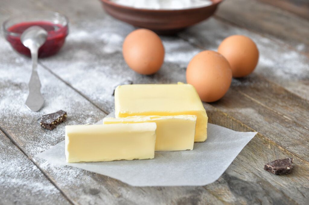bloco de margarina culinária em fatias sobre papel manteiga em uma bancada rústica de madeira polvilhada com farinha, acompanhado de ovos inteiros e uma pequena tigela com calda de frutas vermelhas.