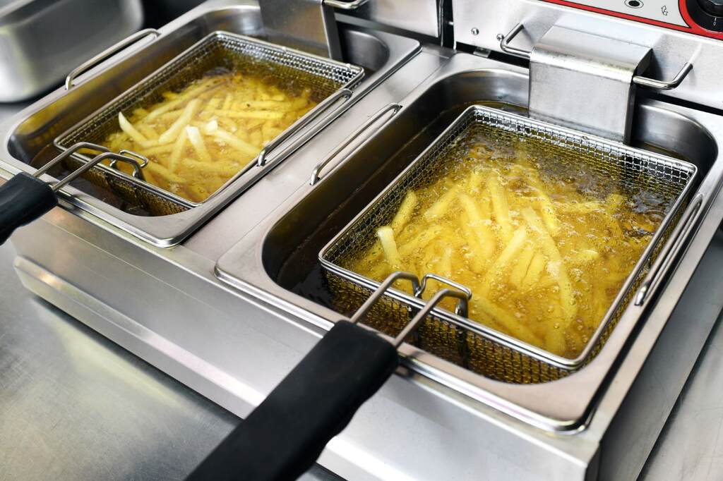 Batatas fritas sendo preparadas em fritadeira de imersão profissional com cestos metálicos. Detalhe do óleo borbulhando em cozinha industrial.