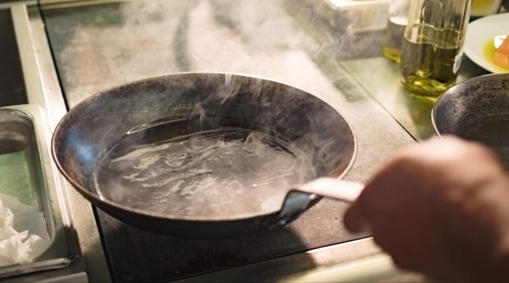 Frigideira de metal em fogo alto com óleo atingindo o ponto de fumaça, soltando vapores brancos em uma cozinha profissional.