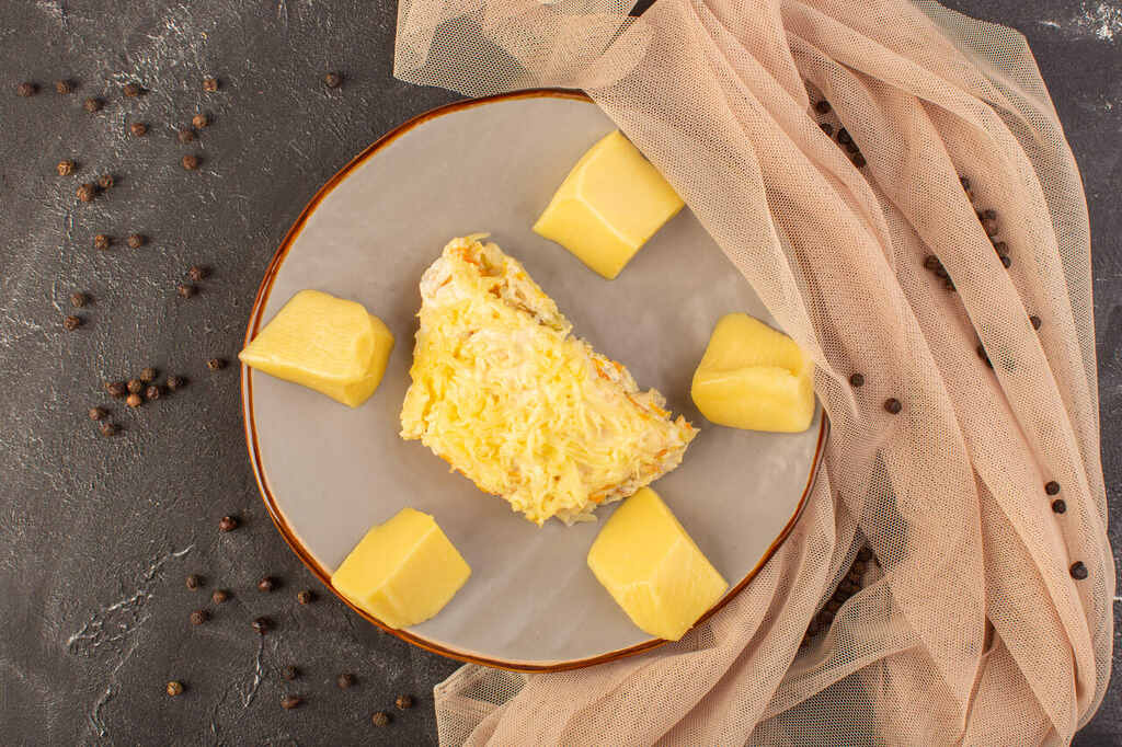 Vista superior de um prato cinza contendo uma fatia de bolo salgado ou torta com queijo ralado, cercada por cinco pedaços de manteiga ou margarina. A composição inclui um tecido bege e grãos de pimenta espalhados sobre uma superfície escura.