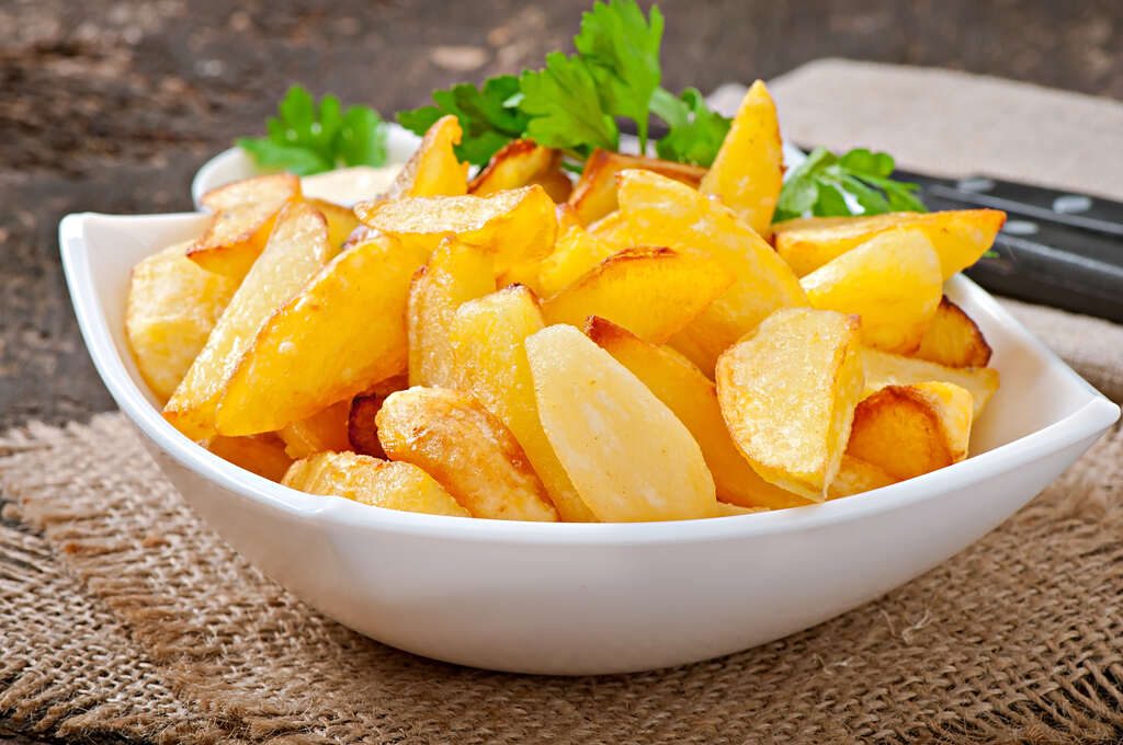 Uma tigela branca de cerâmica repleta de batatas rústicas fritas, bem douradas, decoradas com folhas de salsa fresca. A tigela está posicionada sobre um descanso de juta em uma mesa de madeira escura, transmitindo uma aparência apetitosa.