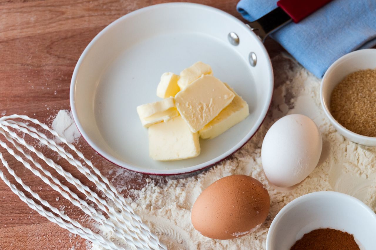 Margarina em pedaços dentro de frigideira branca ao lado de ovos, farinha e açúcar para preparo de receita