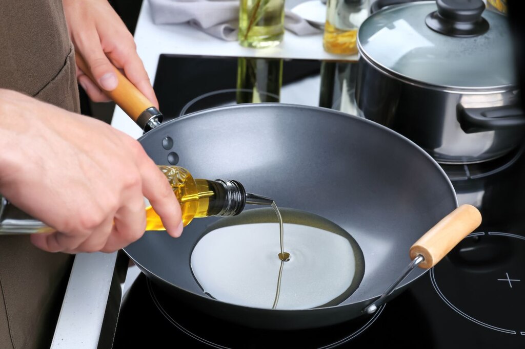 Pessoa despejando óleo vegetal de uma garrafa de vidro em uma panela wok sobre um fogão de indução moderno. A imagem foca na ação de cozinhar com gordura vegetal em uma cozinha profissional.