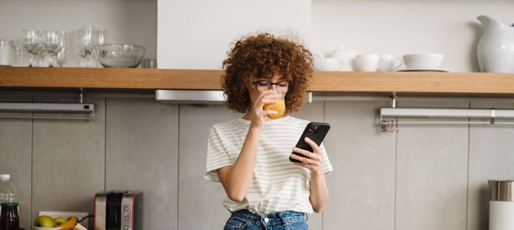 Mulher na cozinha bebendo suco e usando celular durante preparo de alimentos