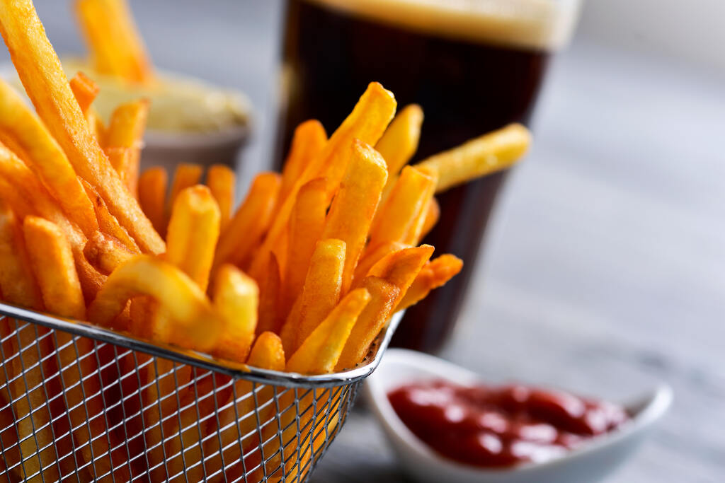 Porção de batata frita crocante servida em cestinha metálica com ketchup e bebida ao fundo