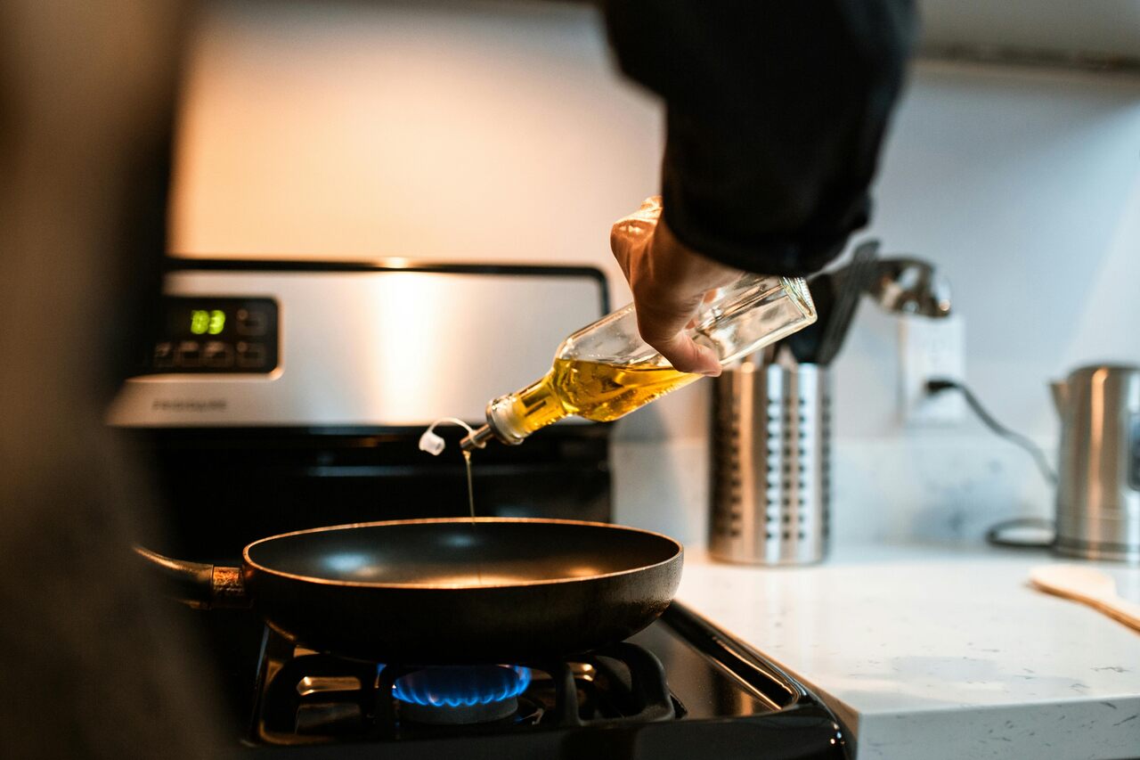 Pessoa despejando óleo vegetal em frigideira sobre fogão aceso durante preparo de alimentos