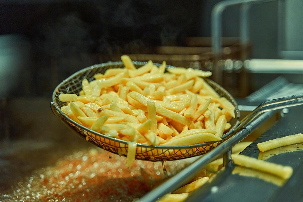 Batatas fritas sendo retiradas da fritadeira industrial com escorredor metálico, destacando o preparo em óleo quente.
