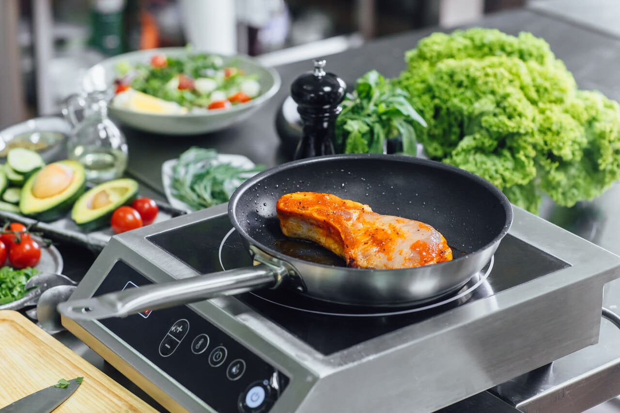 Carne sendo grelhada na frigideira com legumes frescos ao fundo em cozinha moderna