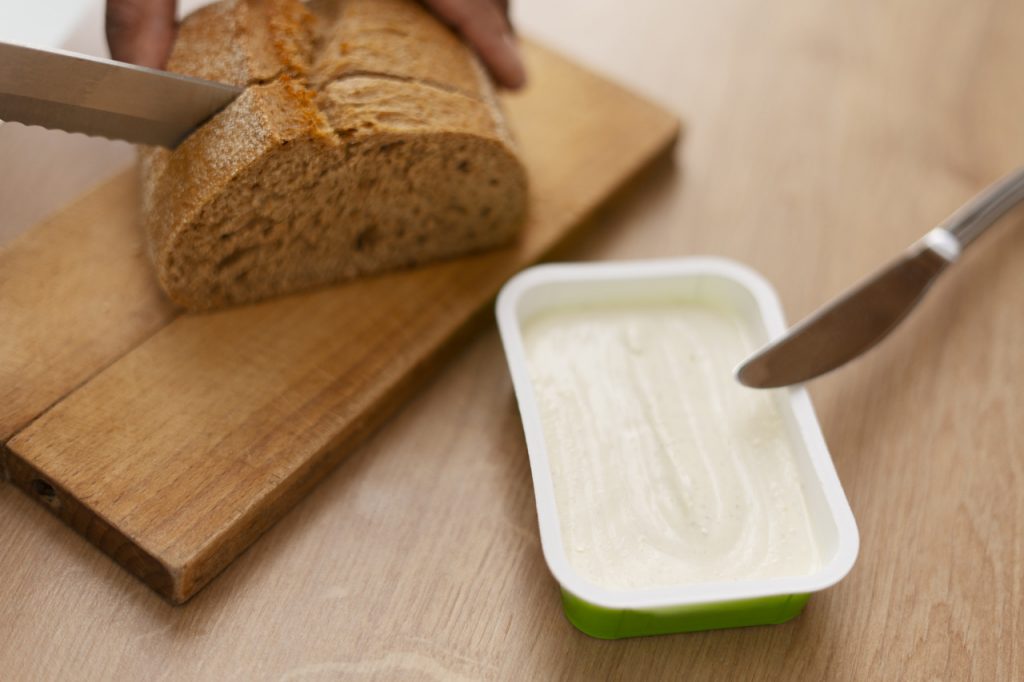 Margarina em pote ao lado de pão sendo cortado sobre tábua de madeira na cozinha