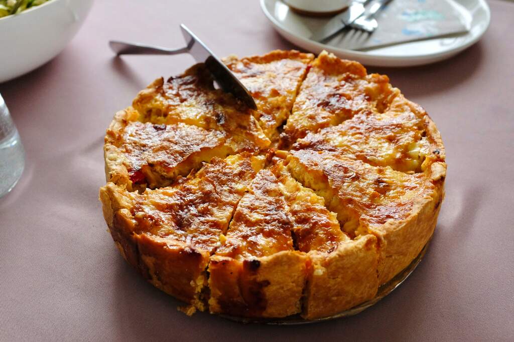 Torta salgada assada e fatiada sobre a mesa, com cobertura gratinada dourada e utensílio para servir ao lado.