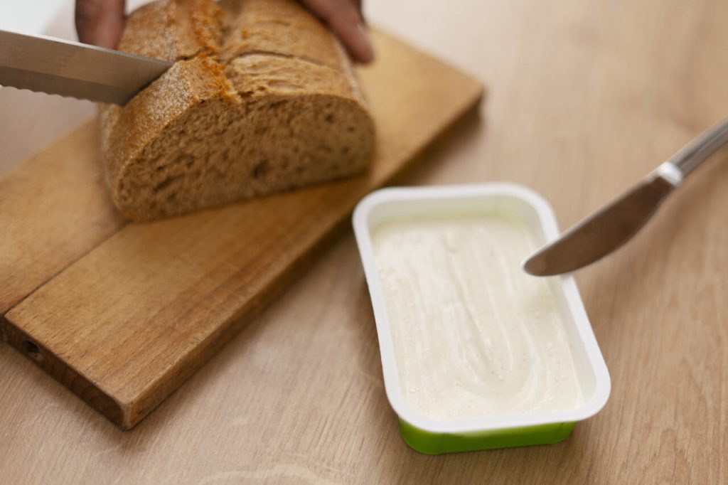 Pão sendo cortado ao lado de pote de margarina representando uso no dia a dia