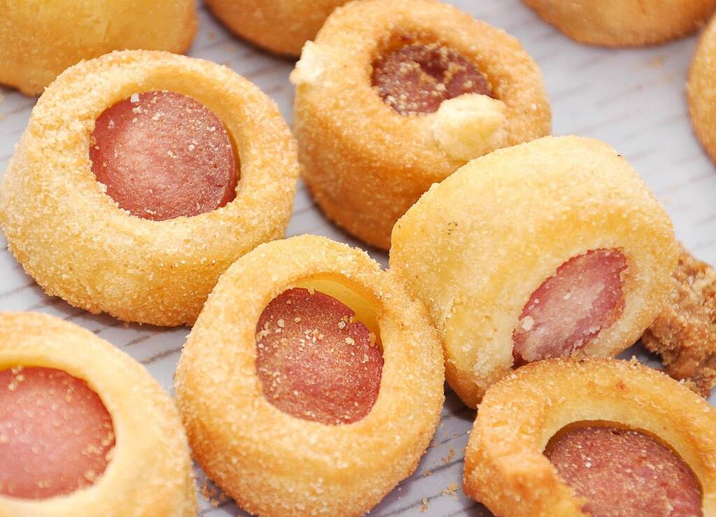 Mini salgados empanados recheados com salsicha, fritos e servidos em close-up, destacando a textura crocante.