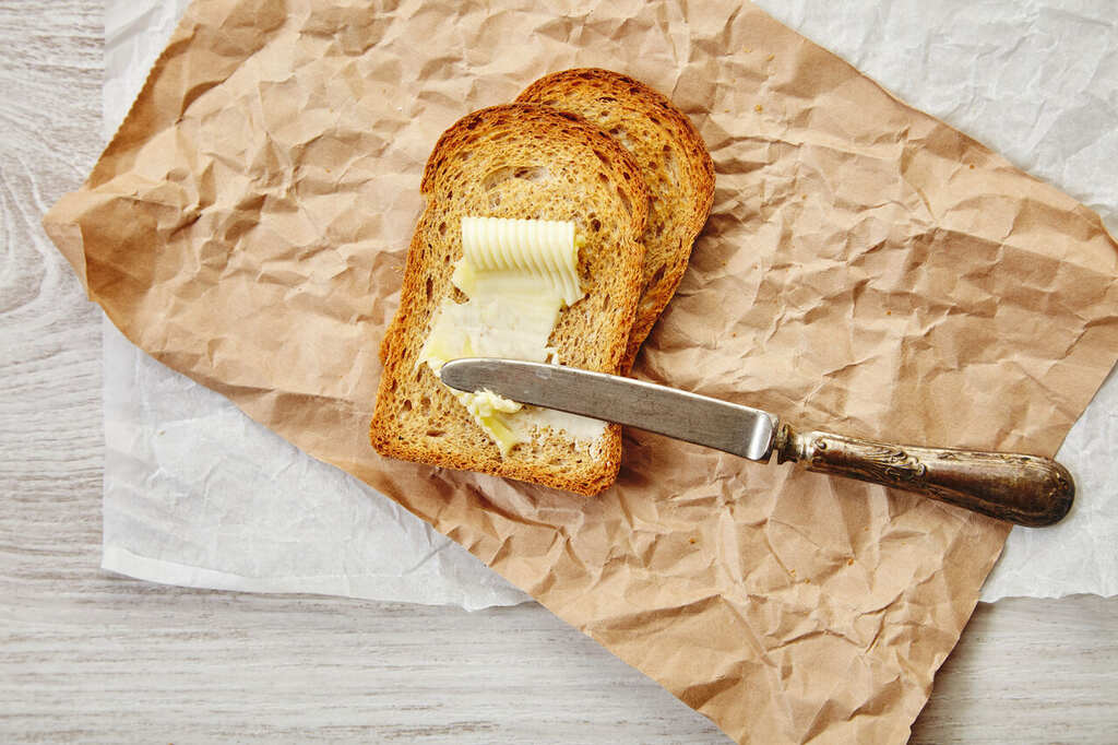 Fatias de pão torrado com margarina sendo espalhada com faca sobre papel kraft em mesa clara